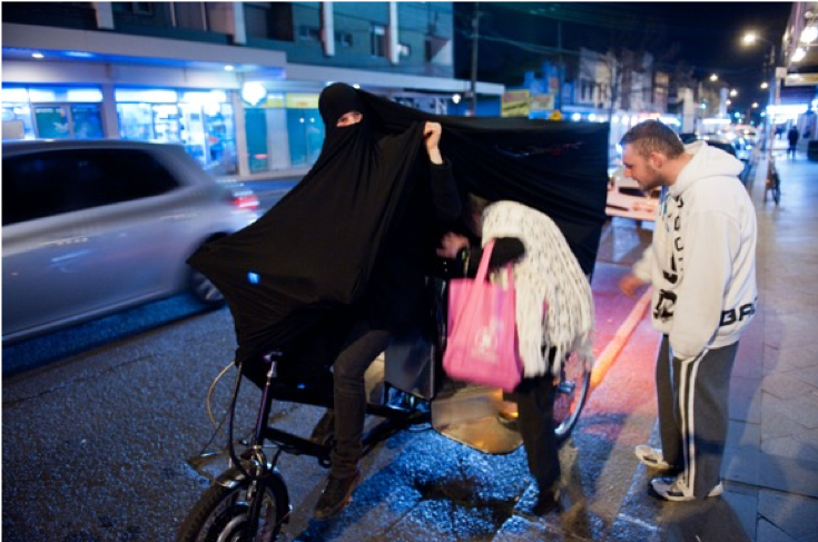 Woman in black burqa driving bicycle and picking up passengers.