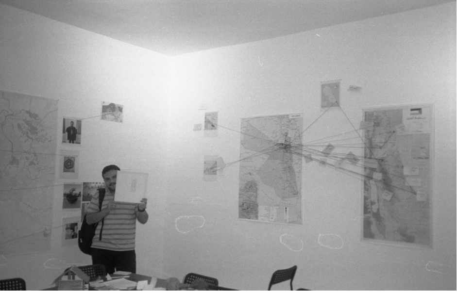 A man wearing a striped shirt and a backpack holds up a white square next to his face. He is standing in the workshop room, with maps and other photos tacked onto the wall behind him.