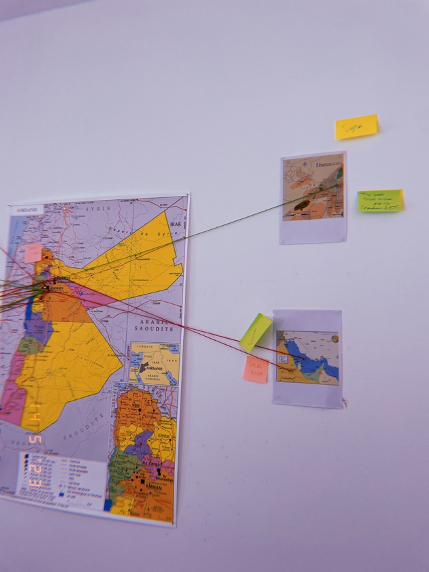 One large and two smaller maps are situated next to each other on a wall. Red strings and sticky notes indicate connections between the three locations.